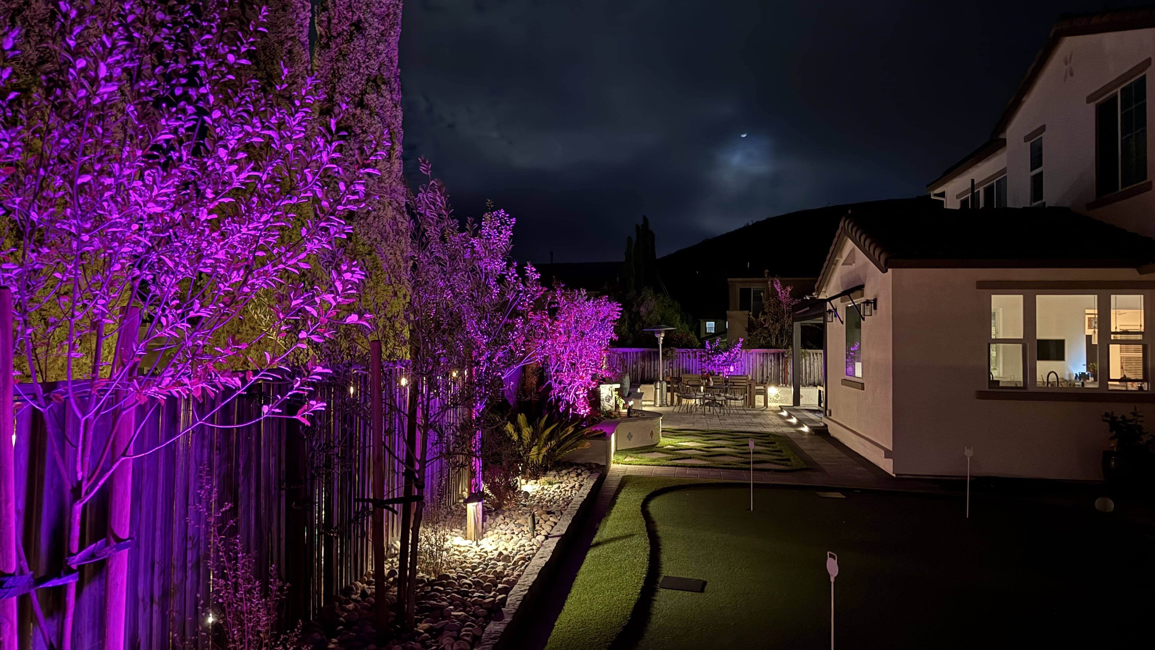 Color-changing garden lights Purple landscape lights illuminate trees beside a backyard putting green, with warm lighting along the planted border and paving.